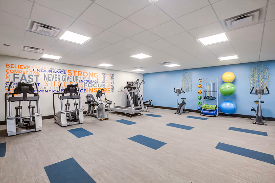 A spacious fitness room with exercise equipment including treadmills, stationary bikes, and elliptical machines. The walls are painted white and blue, with motivational words like 'STRONG', 'SUCCESS', and 'NEVER GIVE UP' displayed on one wall. Exercise balls and other fitness accessories are neatly arranged on a rack against the blue wall. The floor has a light wood pattern with blue rectangular accents.
