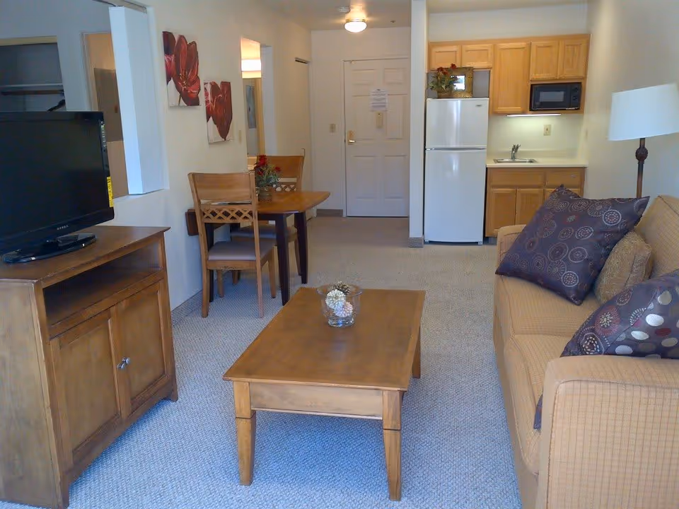 A cozy senior living apartment interior featuring a beige sofa with patterned pillows, a wooden coffee table with a decorative centerpiece, a wooden TV stand with a flat-screen television, a small dining table with two chairs, and a kitchenette with a refrigerator, microwave, sink, and wooden cabinets. The entrance door is visible in the background.
