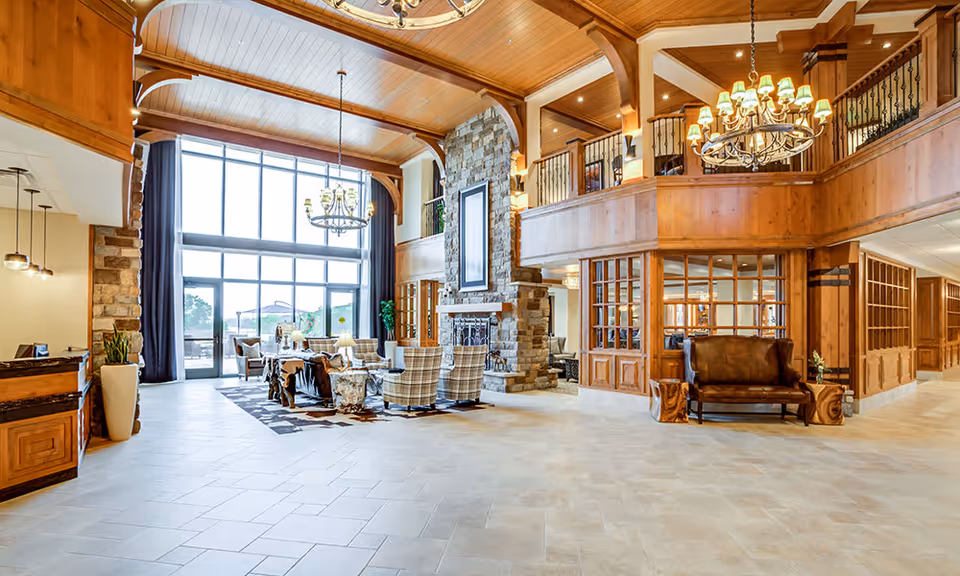 Spacious senior living facility common area with high wooden ceilings, large windows letting in natural light, a stone fireplace, chandeliers, and comfortable seating including armchairs and a bench.