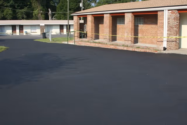 View of a paved parking lot area in front of a single-story brick building with multiple doorways, and another similar building in the background. Yellow caution tape is stretched across the front of the brick building.