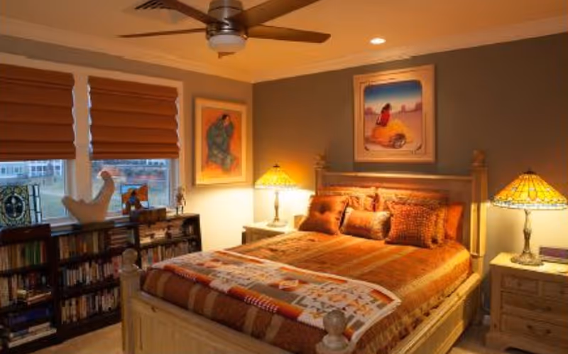 Cozy bedroom with a wooden bed covered in a warm-toned quilt and pillows, flanked by two bedside tables with stained-glass lamps, a bookshelf under the windows, and a ceiling fan.