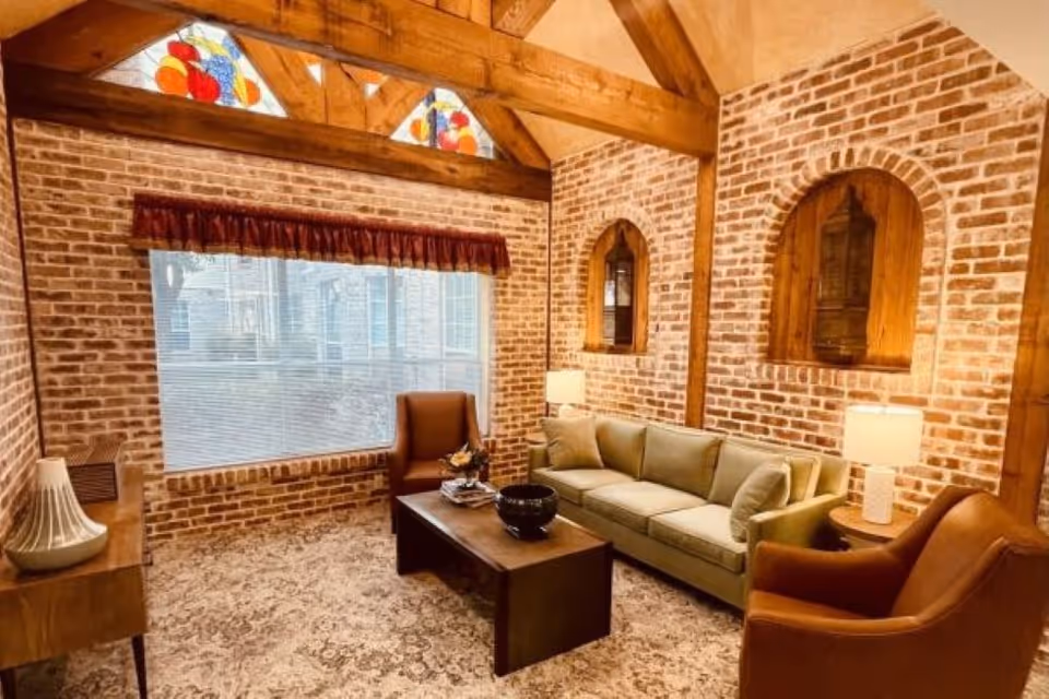 Cozy living room with exposed brick walls, wooden beams, a sofa, armchairs, coffee table, and stained-glass windows.