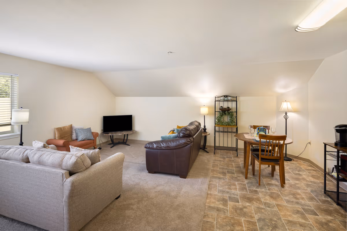 A cozy senior living community room featuring a beige sofa, a brown leather sofa, and an orange armchair arranged around a small TV on a stand. The room has a mix of carpet and tiled flooring, with a small wooden dining table and two chairs on the tiled side. There are two floor lamps providing warm lighting, a decorative metal shelving unit with plants and bowls, and a coffee maker on a small black metal cart.