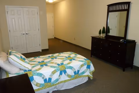 A furnished bedroom in a senior living facility with a quilted bed and dresser.