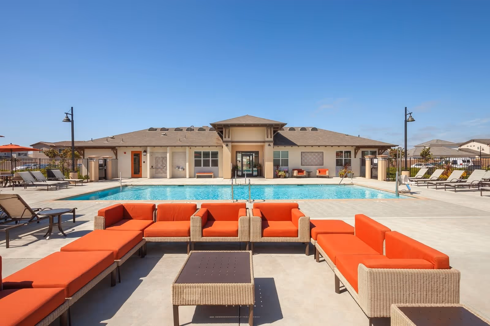 Outdoor pool area at Villa del Sol with a large swimming pool surrounded by lounge chairs and a seating area with orange cushioned wicker sofas and a table. The building in the background has a covered entrance and windows, under a clear blue sky.