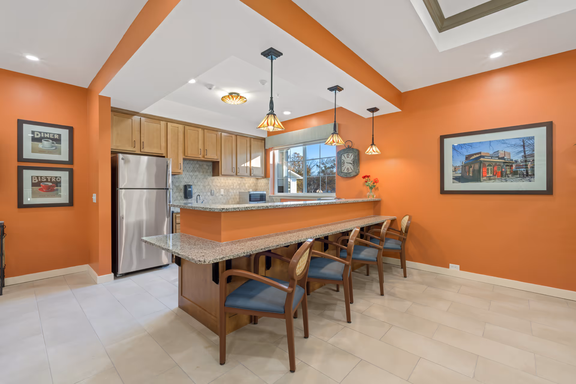 Interior view of a kitchen area with orange walls, wooden cabinets, a stainless steel refrigerator, and a granite countertop bar with four wooden chairs. The room is decorated with framed pictures on the walls, a clock, and pendant lights hanging from the ceiling.