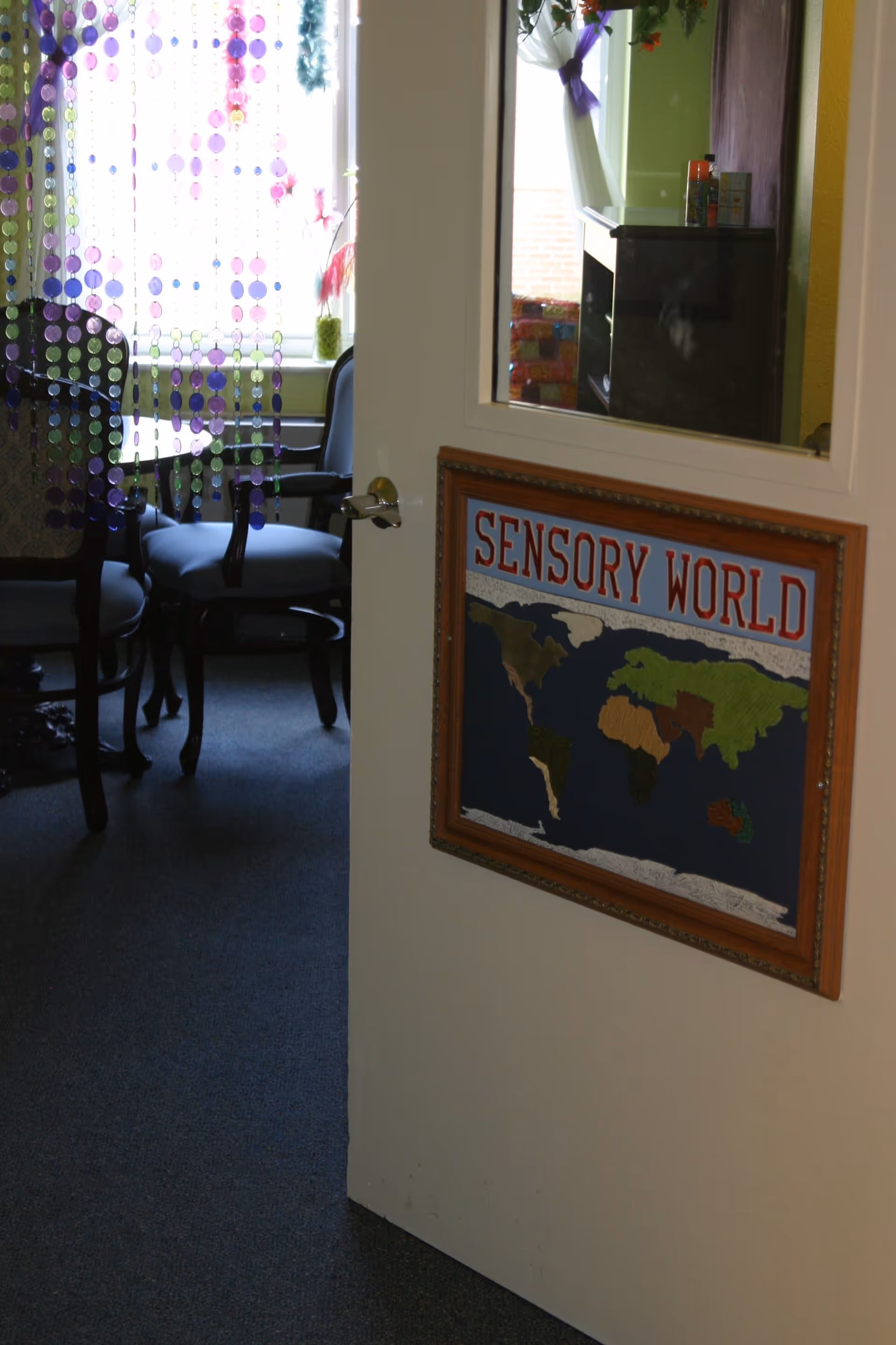 Partially open door with a framed sign reading 'SENSORY WORLD' and a world map below the text. Through the doorway, a room with chairs around a table is visible, with a window covered by hanging purple and pink bead curtains letting in natural light.
