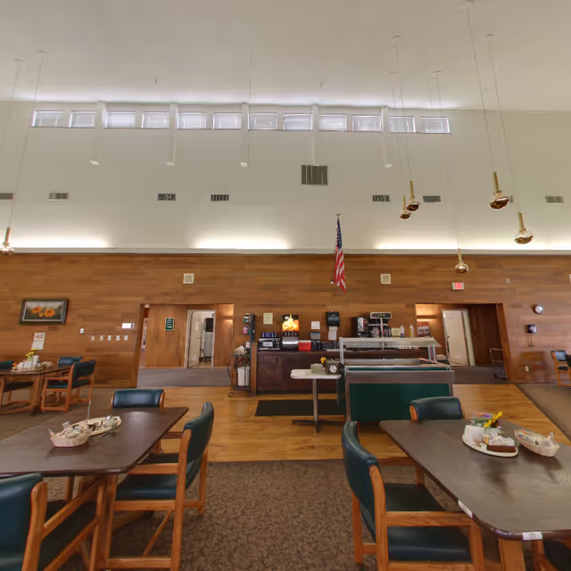 Interior view of a dining area in a retirement community with wooden tables and green cushioned chairs. The room features a high ceiling with small windows near the top, pendant lights hanging down, and a wooden accent wall with an American flag. There is a counter area with coffee machines and other beverage dispensers.