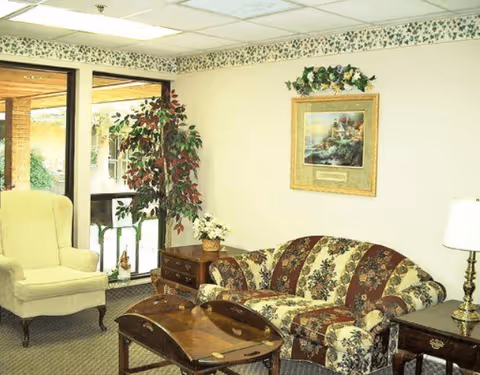 A cozy living room area with a floral patterned sofa, a beige armchair, two wooden side tables with lamps, a wooden coffee table, a potted plant, and a framed painting on the wall. There is a large window with a view of an outdoor area.