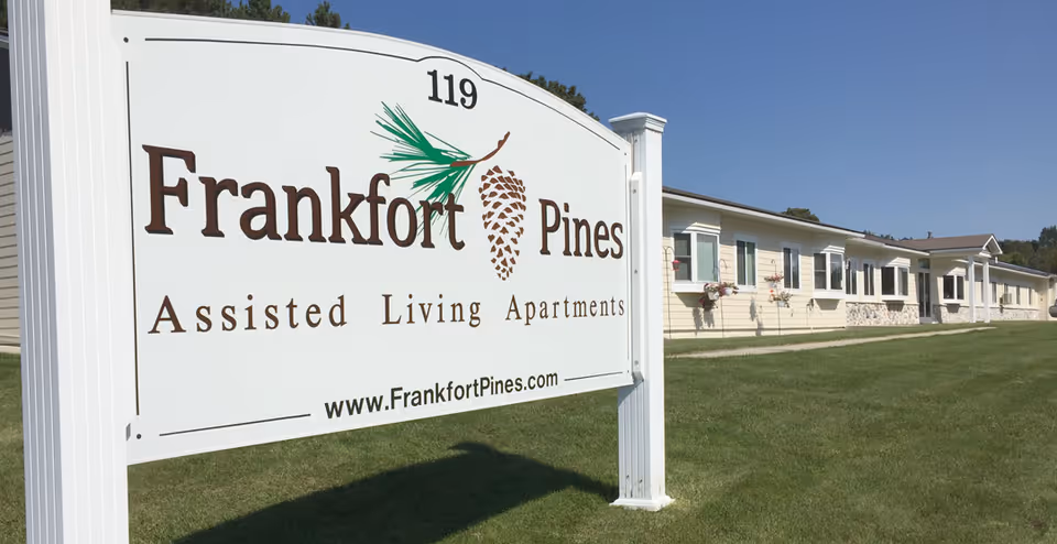A large white sign with brown text reading 'Frankfort Pines Assisted Living Apartments' with a pine cone and pine needle graphic, positioned on a grassy lawn in front of a long, single-story beige building under a clear blue sky.
