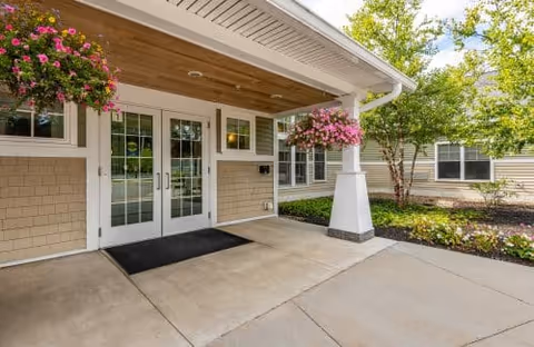 Entrance to a building with double glass doors, beige siding, and a covered porch area. Two hanging flower baskets with pink flowers are on either side of the entrance. There is a black doormat in front of the doors and landscaping with green trees and shrubs nearby.