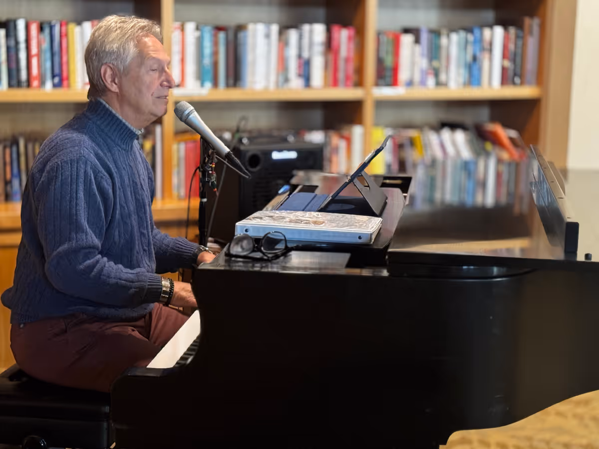 An elderly man wearing a blue sweater is sitting at a black grand piano, singing into a microphone. Behind him is a bookshelf filled with books. On the piano are a pair of glasses, a binder, and a tablet on a stand.