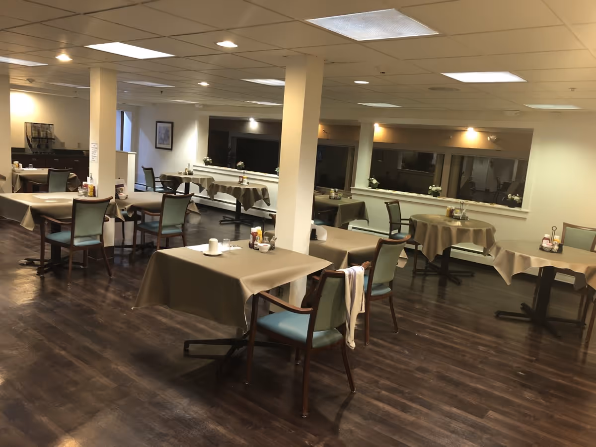 Interior view of a senior living facility dining room with multiple tables covered with beige tablecloths and chairs with green cushions. The room has dark wooden flooring, white walls, ceiling lights, and a beverage dispenser in the background. Some tables have condiments and napkins placed on them.