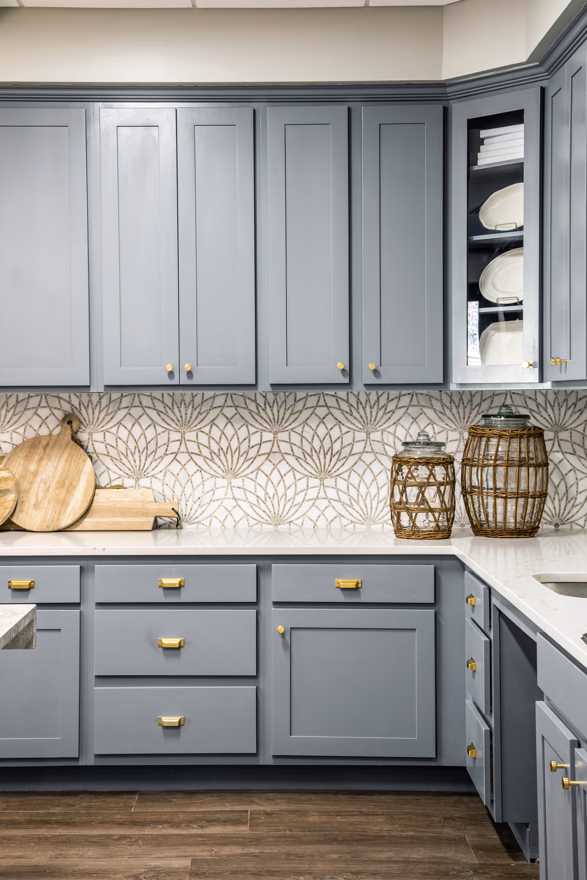 Light gray kitchen cabinets and drawers with brass hardware, a white countertop, patterned tile backsplash, wooden cutting boards and decorative glass jars.