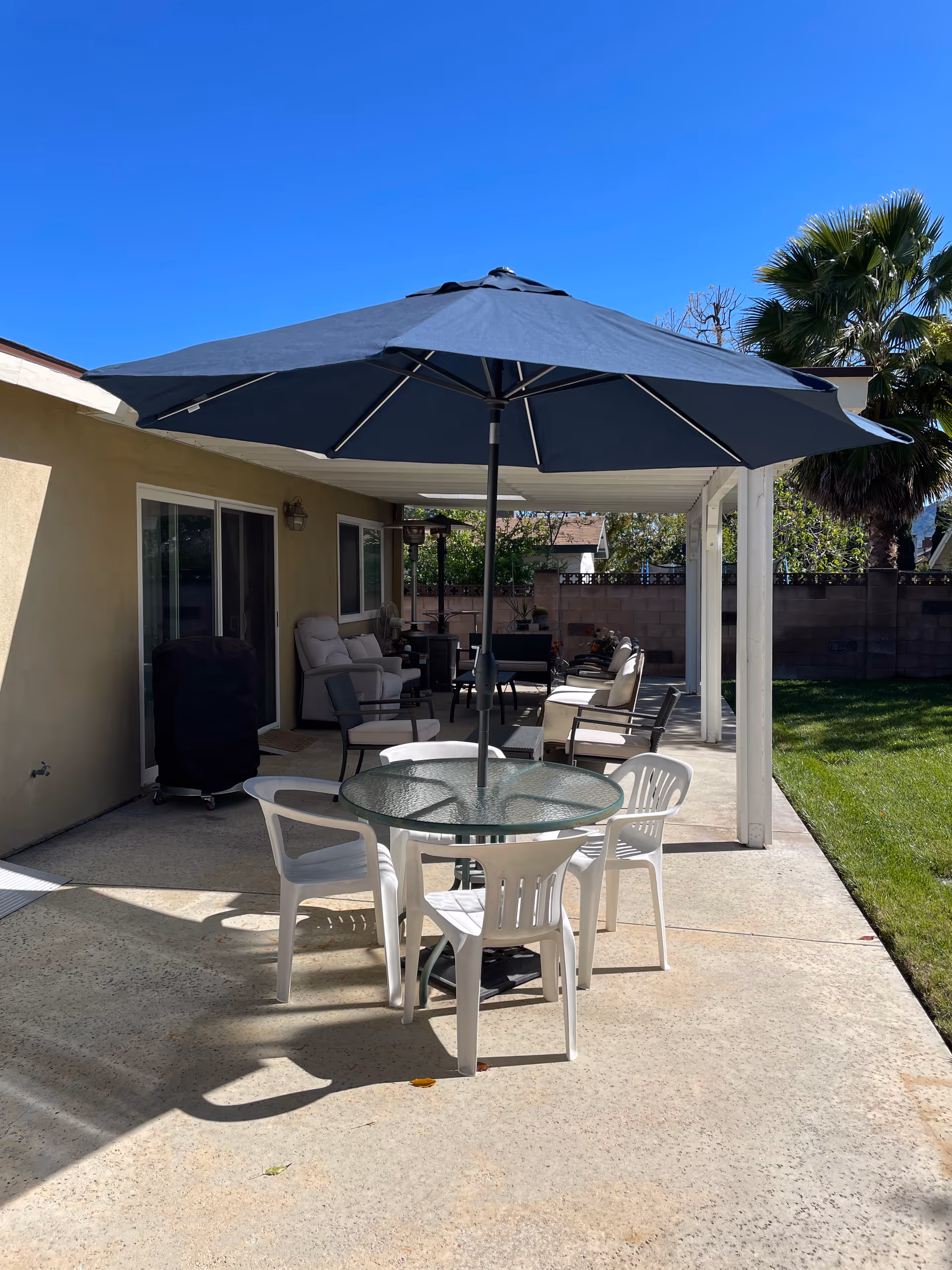 Outdoor patio area with a round glass table and four white plastic chairs under a large dark blue umbrella. The patio is adjacent to a beige building with sliding glass doors and several cushioned chairs arranged under a covered section. There is a green lawn and a palm tree visible on the right side under a clear blue sky.