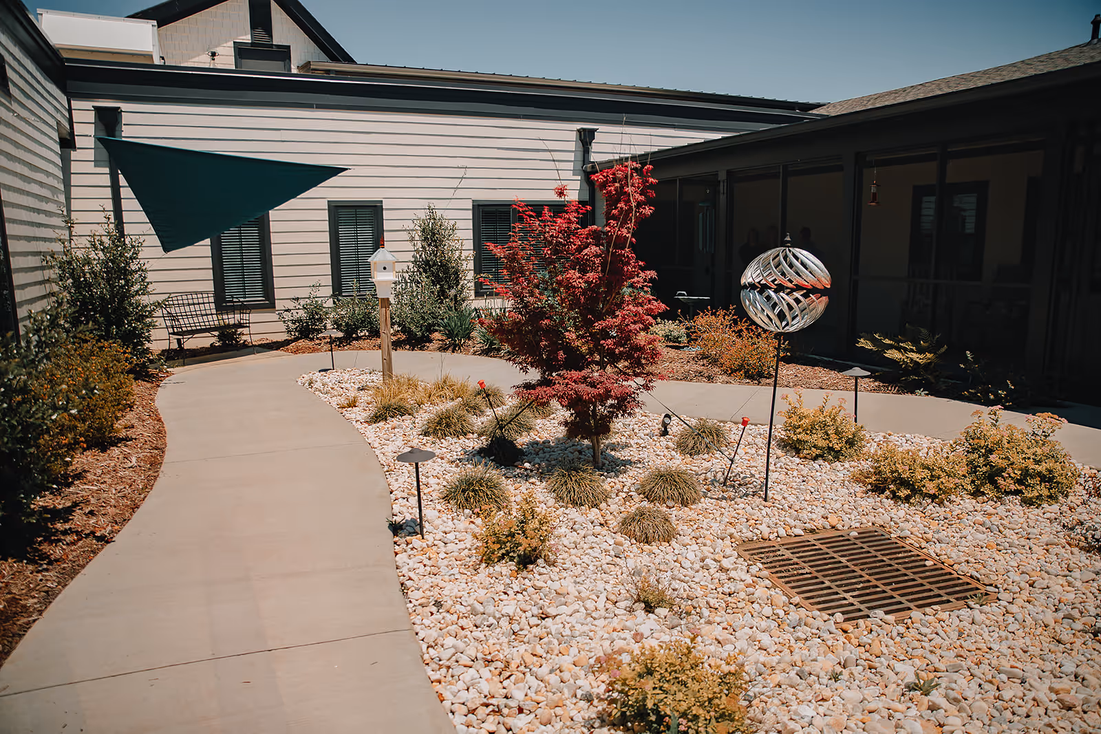 Outdoor courtyard area with a curved concrete pathway surrounded by landscaping featuring small bushes, a red-leafed tree, white decorative rocks, and a metallic garden ornament. The courtyard is bordered by light-colored building walls with windows and a shaded seating area.