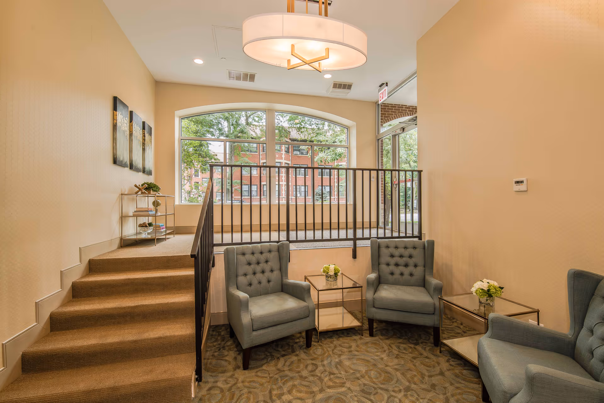 A cozy seating area in a senior living facility with three gray tufted armchairs arranged around two glass-top side tables with small flower arrangements. The area is carpeted with a patterned design and is adjacent to a short staircase leading up to a large window with a view of trees and a brick building outside. The walls are painted beige, and there is a modern circular ceiling light fixture above.