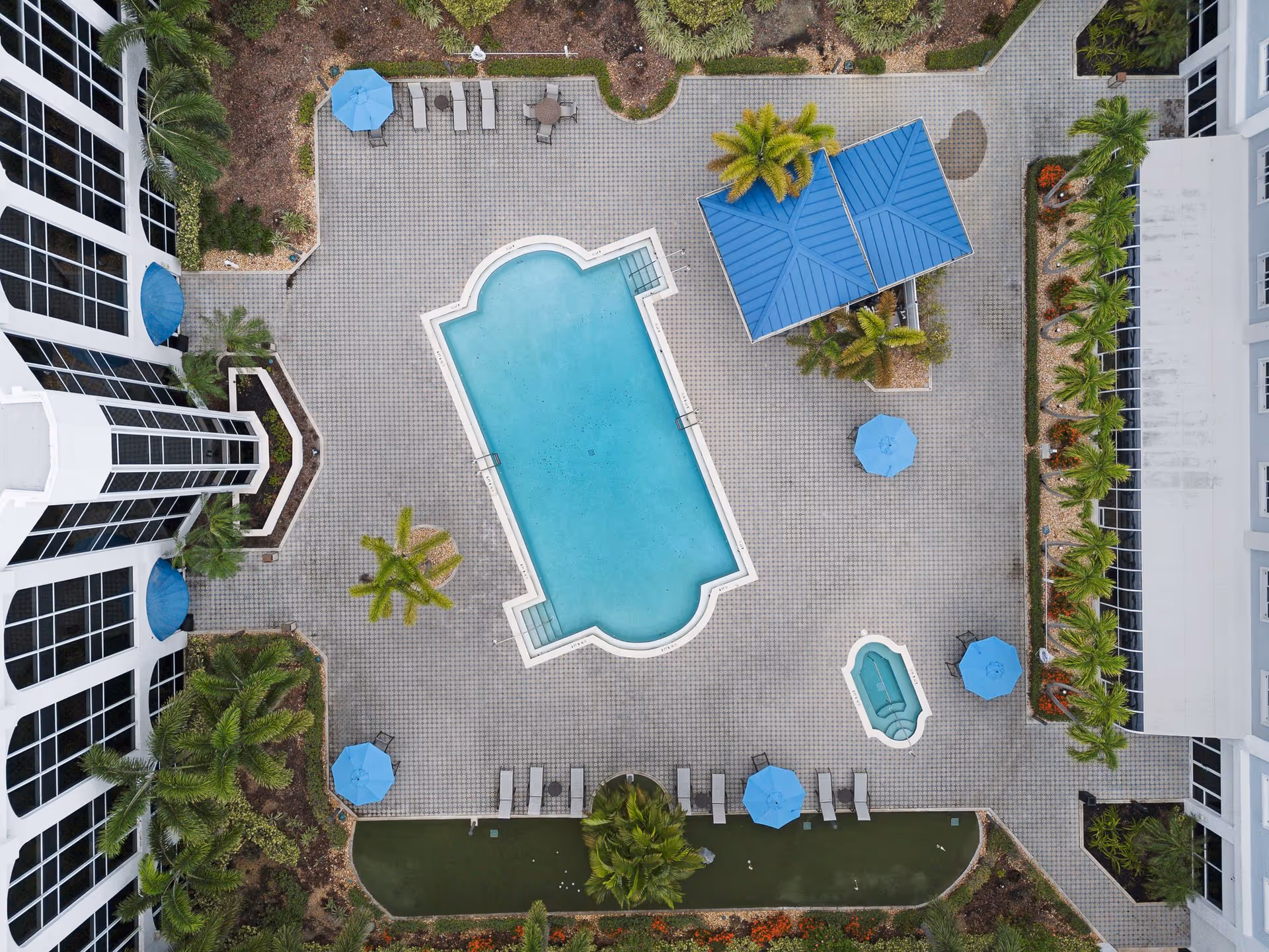 Aerial view of a swimming pool area at The Summit of Fort Myers featuring a large rectangular pool with rounded edges, a smaller hot tub, several blue umbrellas with lounge chairs, palm trees, and surrounding buildings with large windows.