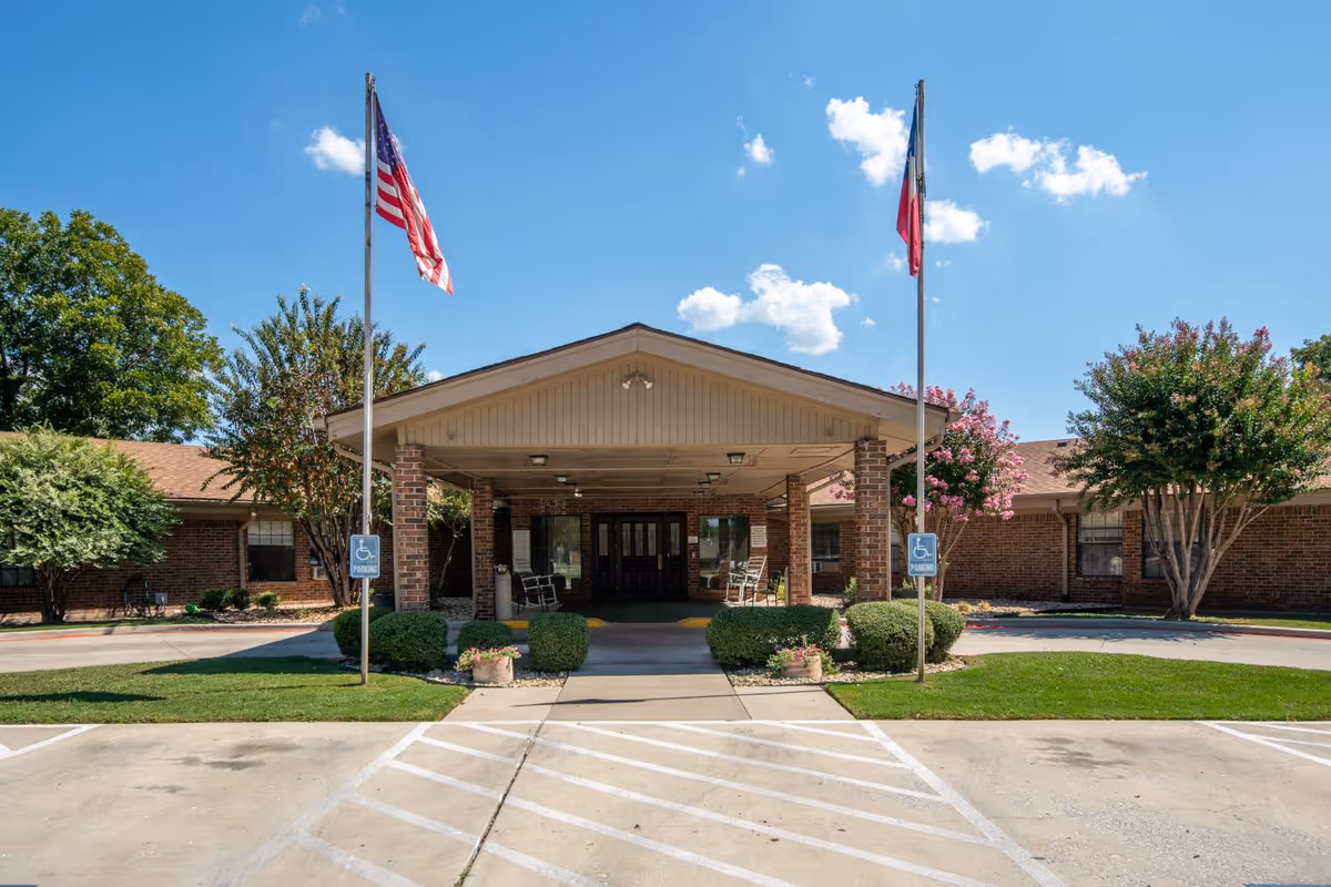 Front entrance of Pecan Tree Rehab and Healthcare Center with a covered drop-off area supported by brick columns, two flagpoles with American and Texas flags, handicap parking signs, and well-maintained landscaping including bushes and trees under a clear blue sky.