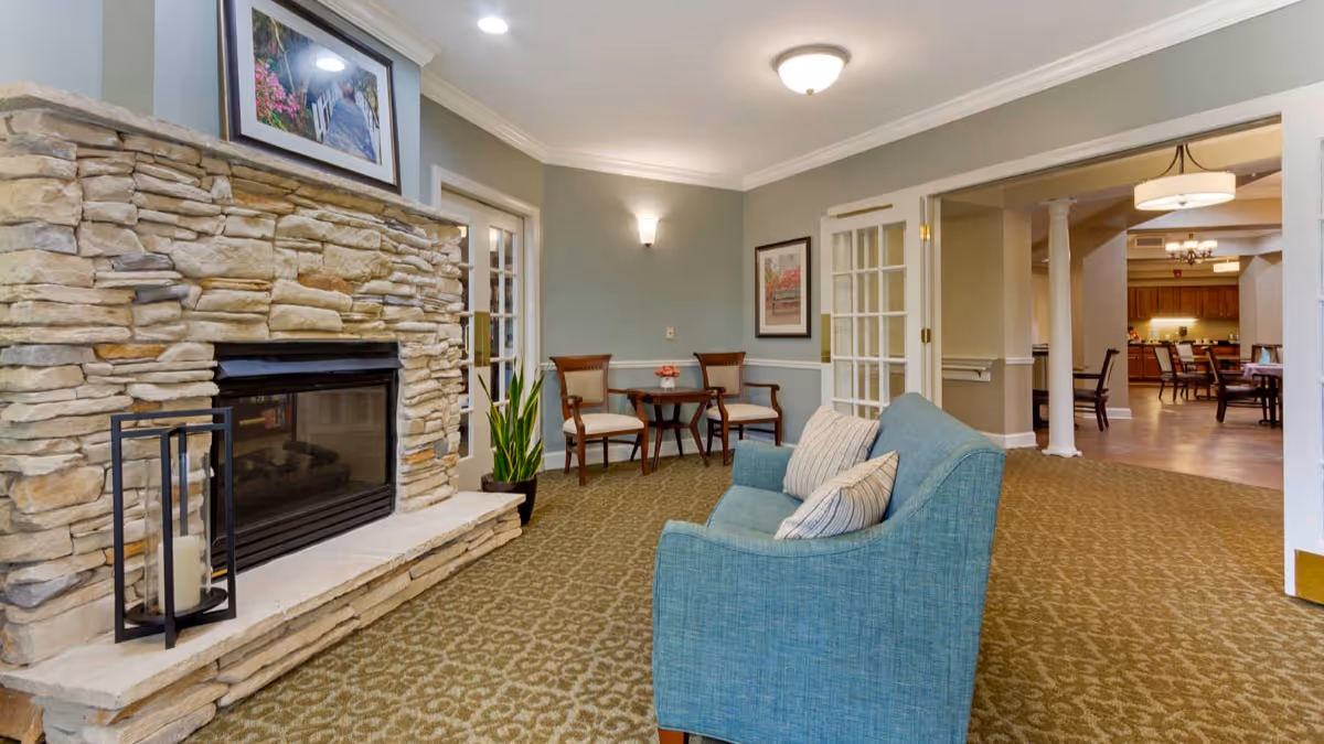 Cozy communal lounge with a stone fireplace, teal sofa, small table and chairs, and a view into a dining area.