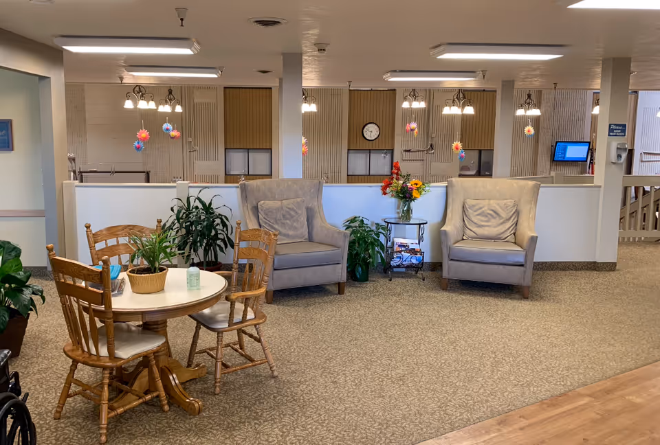 A cozy common area in a senior living facility with a round wooden table surrounded by four wooden chairs, two beige cushioned armchairs with pillows, a small side table with a vase of colorful flowers, several potted plants, and hanging colorful flower decorations. The background shows a clock on the wall and windows with blinds.
