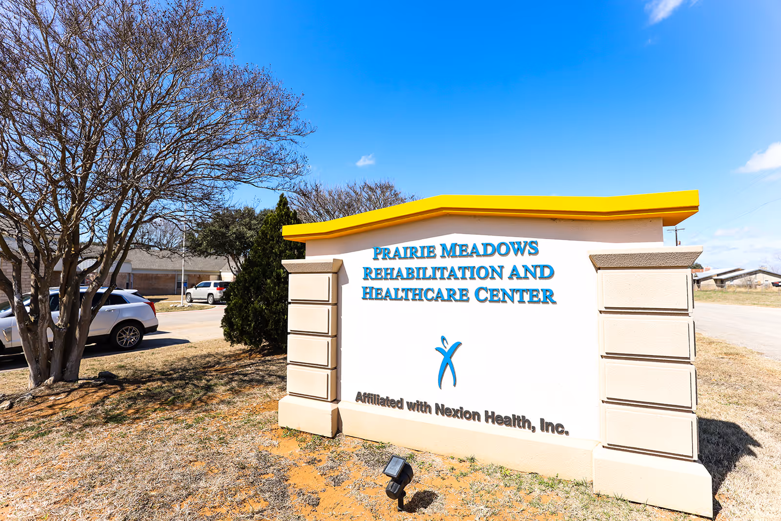Large outdoor sign reading 'Prairie Meadows Rehabilitation and Healthcare Center' with cars, trees, and a blue sky in the background.