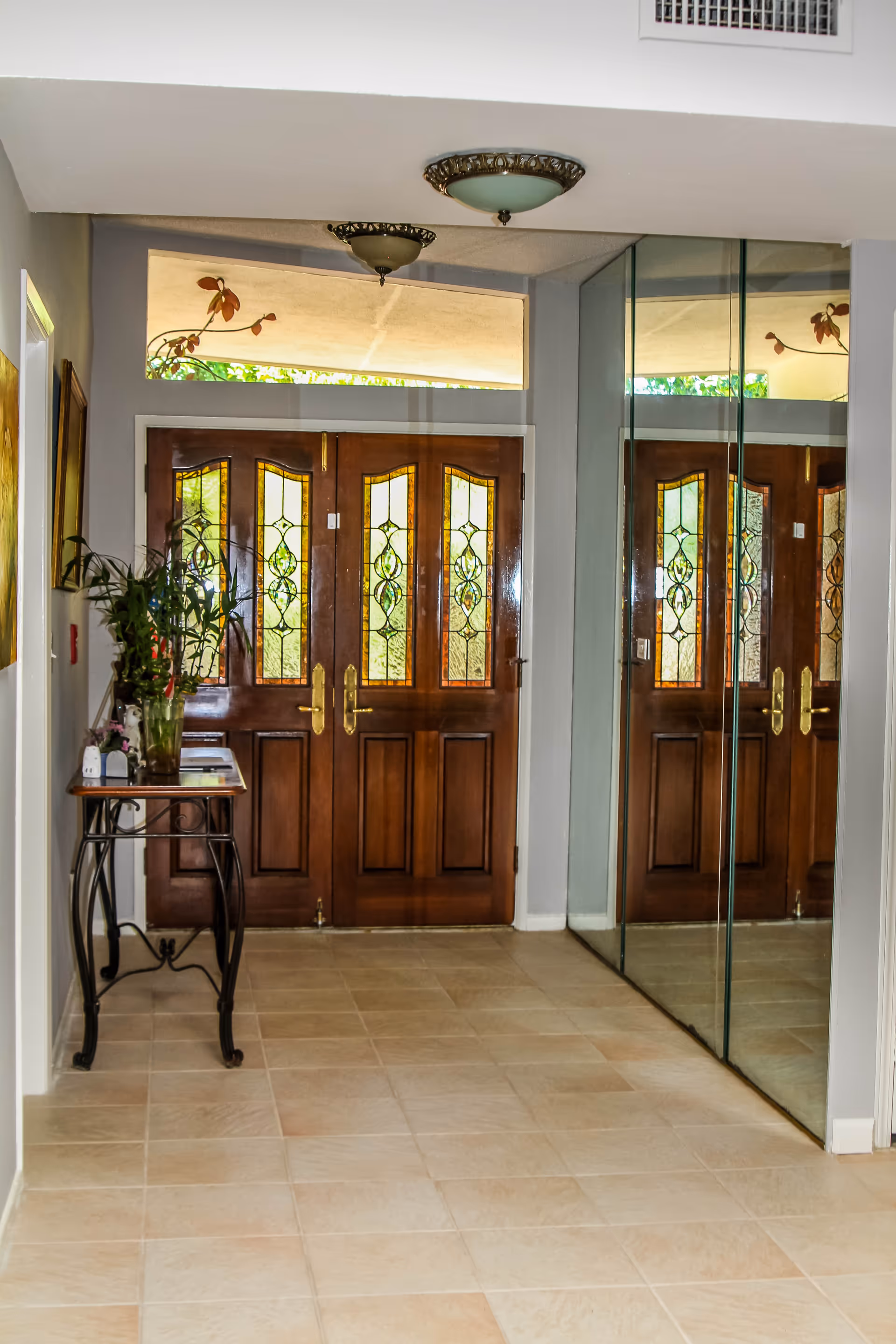Entrance area with double wooden doors featuring decorative glass panels. To the left, there is a small table with a plant and some items on it. On the right side, there is a large mirror reflecting the doors and part of the room. The floor is tiled and the walls are painted light gray.