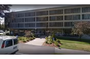 Exterior view of a multi-story building with large windows and a landscaped entrance pathway. There are bushes and small trees in front of the building, and a white vehicle is parked on the left side.