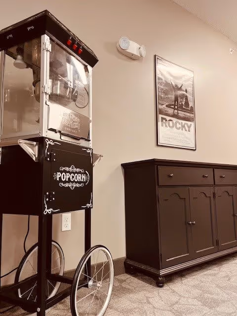 A vintage-style popcorn machine on wheels next to a dark wooden cabinet with drawers and doors, set against a beige wall with a framed Rocky movie poster hanging above.