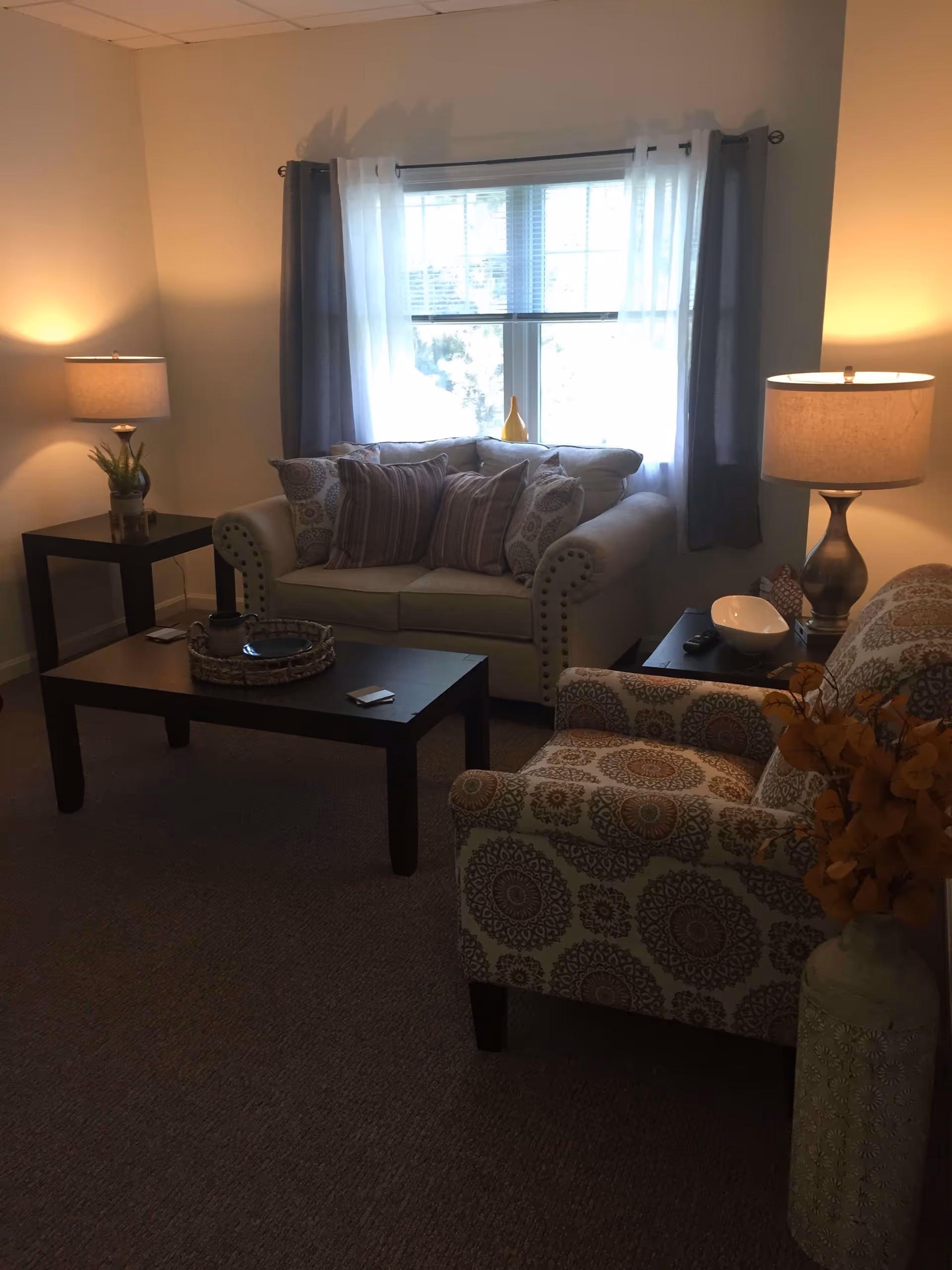 A cozy living room with a beige loveseat adorned with multiple patterned pillows, a patterned armchair, two side tables each with a lamp, a dark wooden coffee table with a decorative tray, and a large window with white and gray curtains letting in natural light.