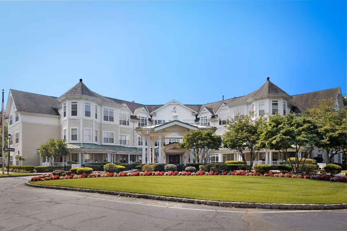 Front exterior of a large three-story white senior living facility with a covered entrance, circular driveway, and manicured lawn and flower beds.