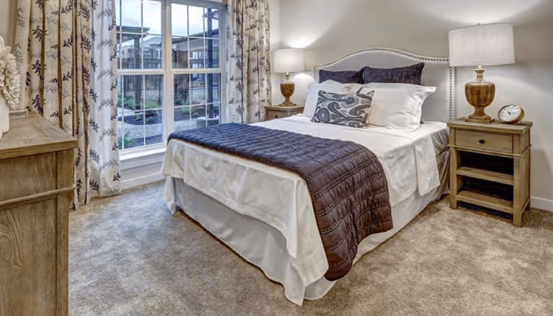 A neatly made bed with white sheets, a dark purple quilted blanket, and decorative pillows in a bedroom. The room has beige carpet, two wooden nightstands with matching lamps, and a large window with patterned curtains allowing natural light to enter.