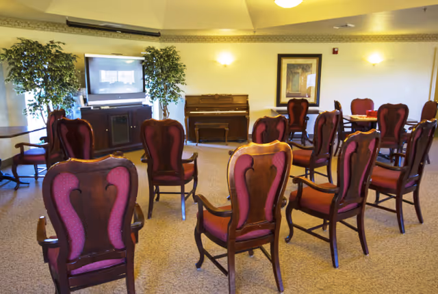 Interior common room with upholstered wooden chairs arranged facing a television and a piano.