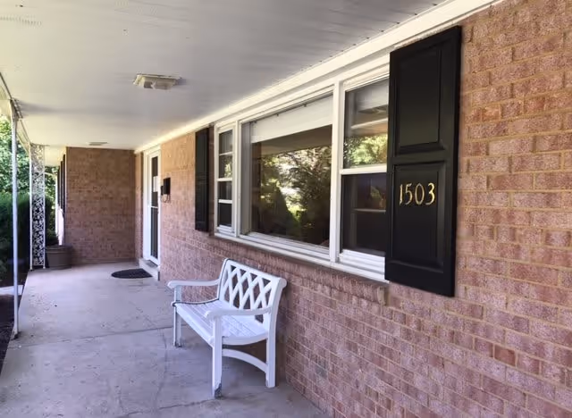 Covered front porch of a brick building with a white bench, black shutters, and the number 1503 displayed on one shutter. The porch has a concrete floor and a white ceiling with a light fixture.