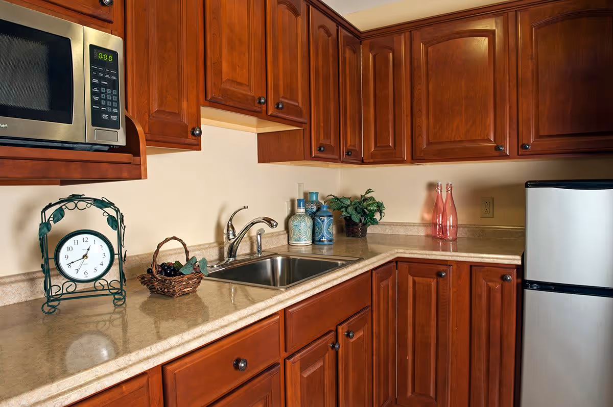 A kitchen area with wooden cabinets, a stainless steel microwave, a small refrigerator, a sink with a faucet, decorative vases, a small basket with grapes, a green decorative clock, and two pink glass bottles on the countertop.