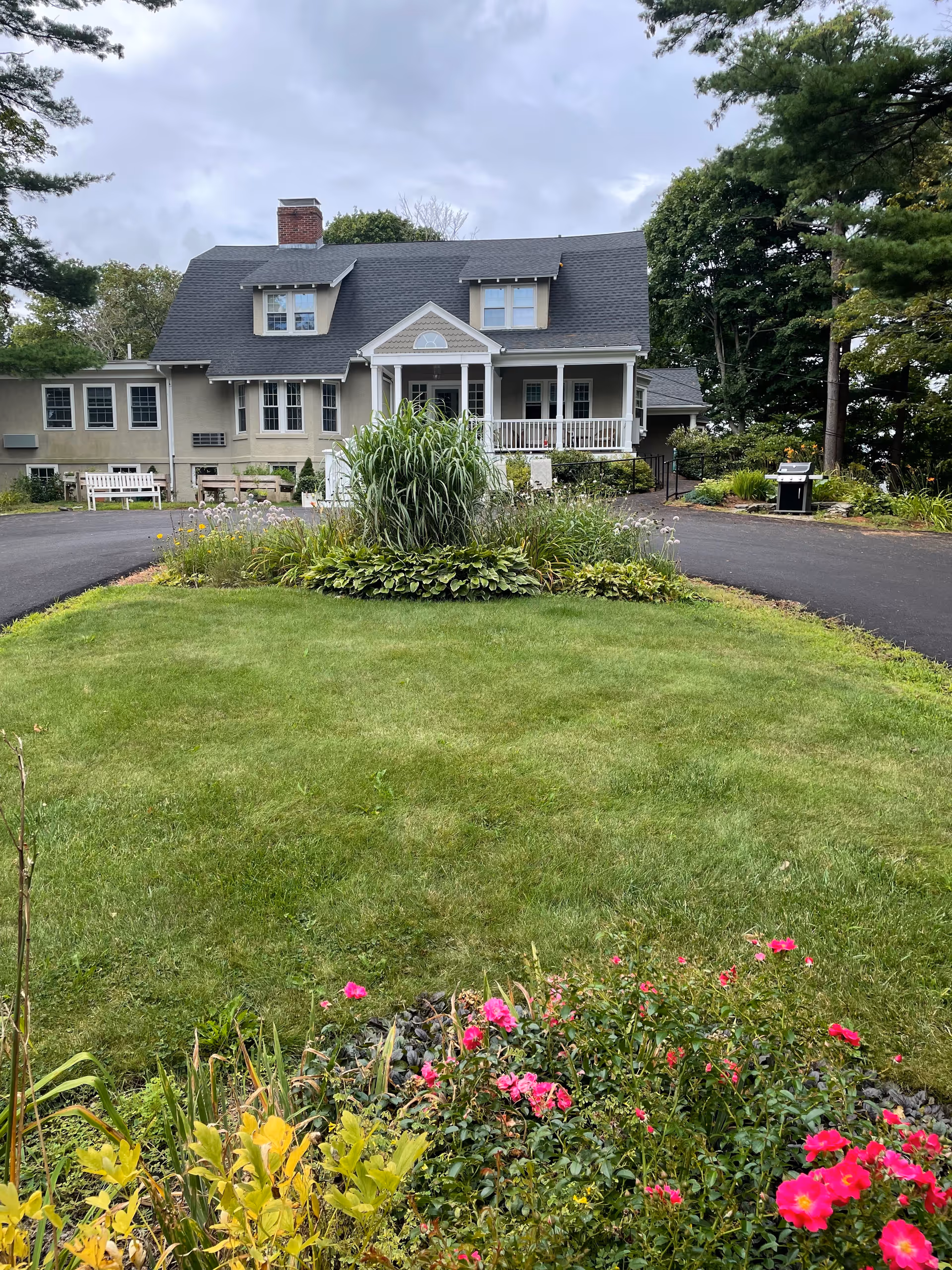 Front exterior of a two-story gray house with a covered porch, circular driveway, and a manicured lawn with flowerbeds.