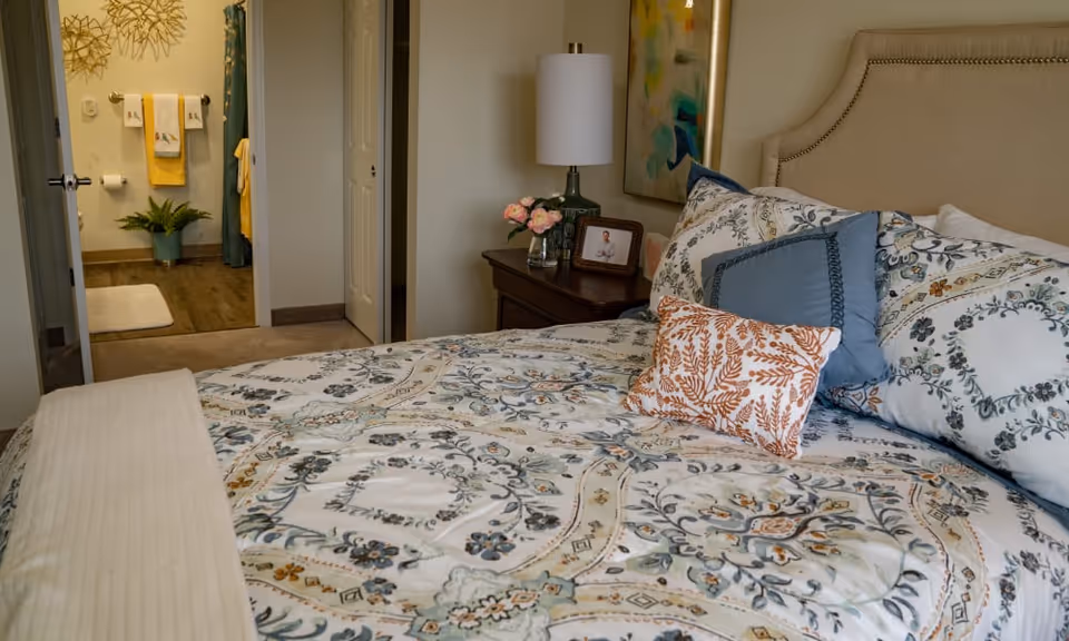 A cozy bedroom with a neatly made bed featuring floral patterned bedding and multiple pillows. A nightstand beside the bed holds a lamp, a small vase with pink flowers, and a framed photo. In the background, an open door reveals a bathroom with towels hanging on the wall and a potted plant on the floor.