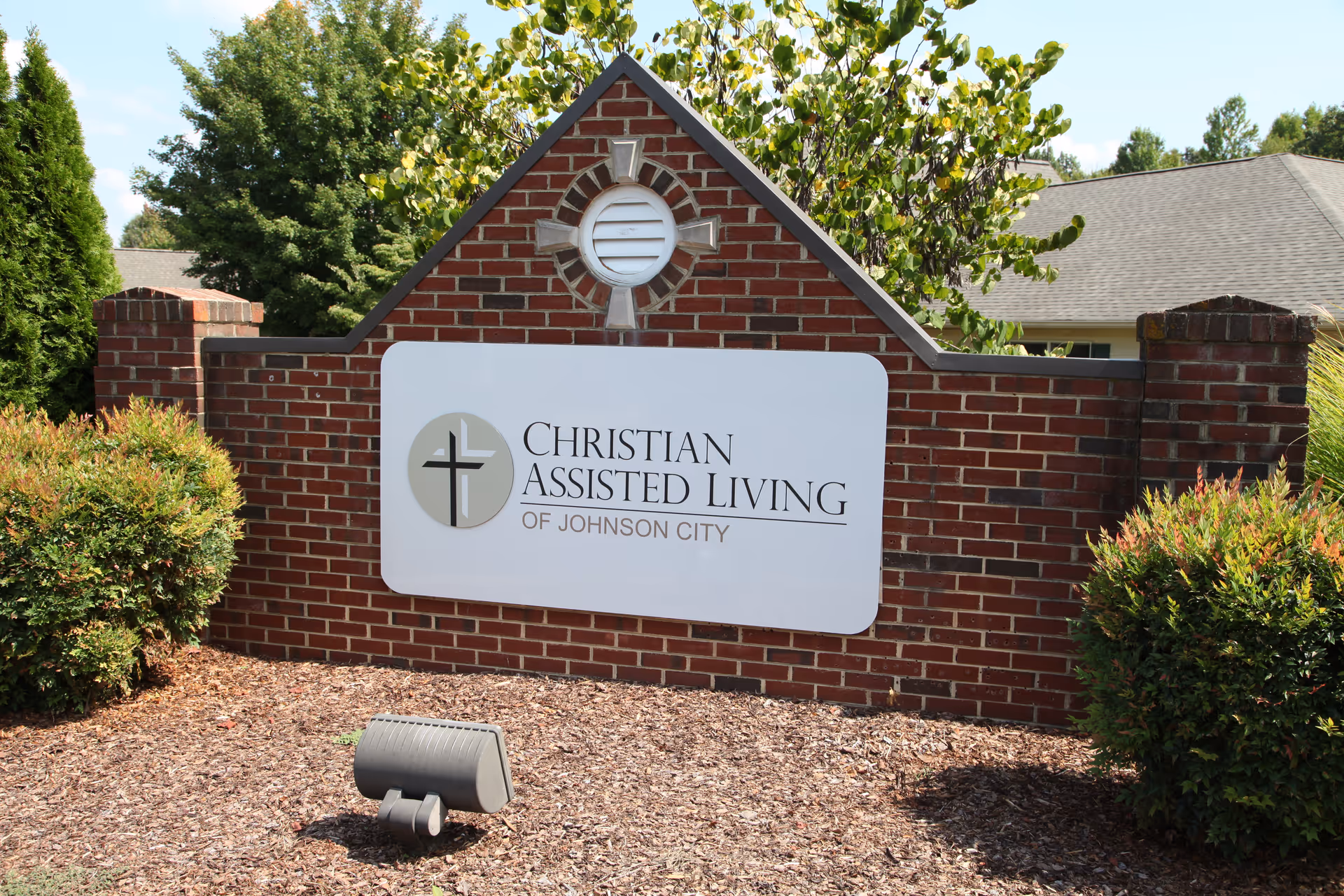 A brick sign for Christian Assisted Living of Johnson City with a cross logo, surrounded by bushes and trees, with a building roof visible in the background.