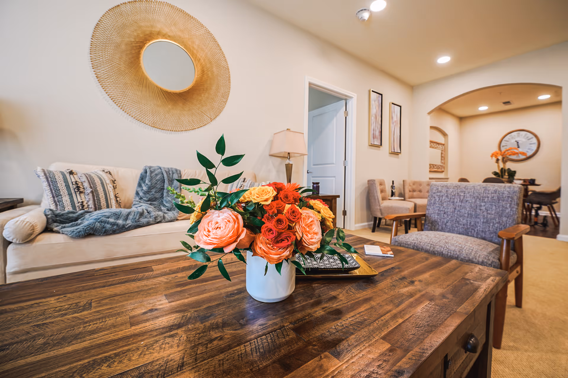 A cozy living room with a wooden coffee table featuring a vase of colorful flowers. Behind the table is a beige sofa with patterned pillows and a blue throw blanket. A round decorative mirror hangs on the wall above the sofa. To the right, there is a gray upholstered chair and a small seating area with two chairs and a table. The room has warm lighting and neutral-colored walls.