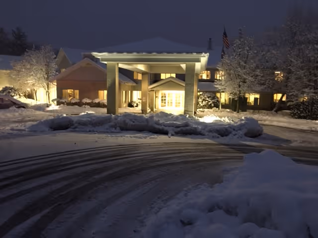 Snow-covered driveway leading to the illuminated front entrance and porte-cochere of a senior living building at night.