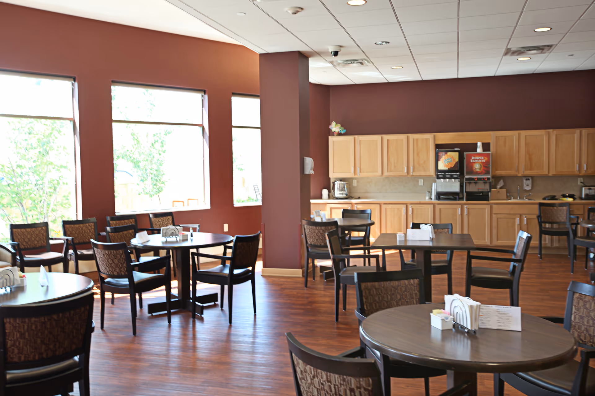 A dining area with several round tables and chairs arranged neatly on a wooden floor. The room has large windows letting in natural light, maroon walls, and a kitchenette area with wooden cabinets and beverage dispensers in the background.