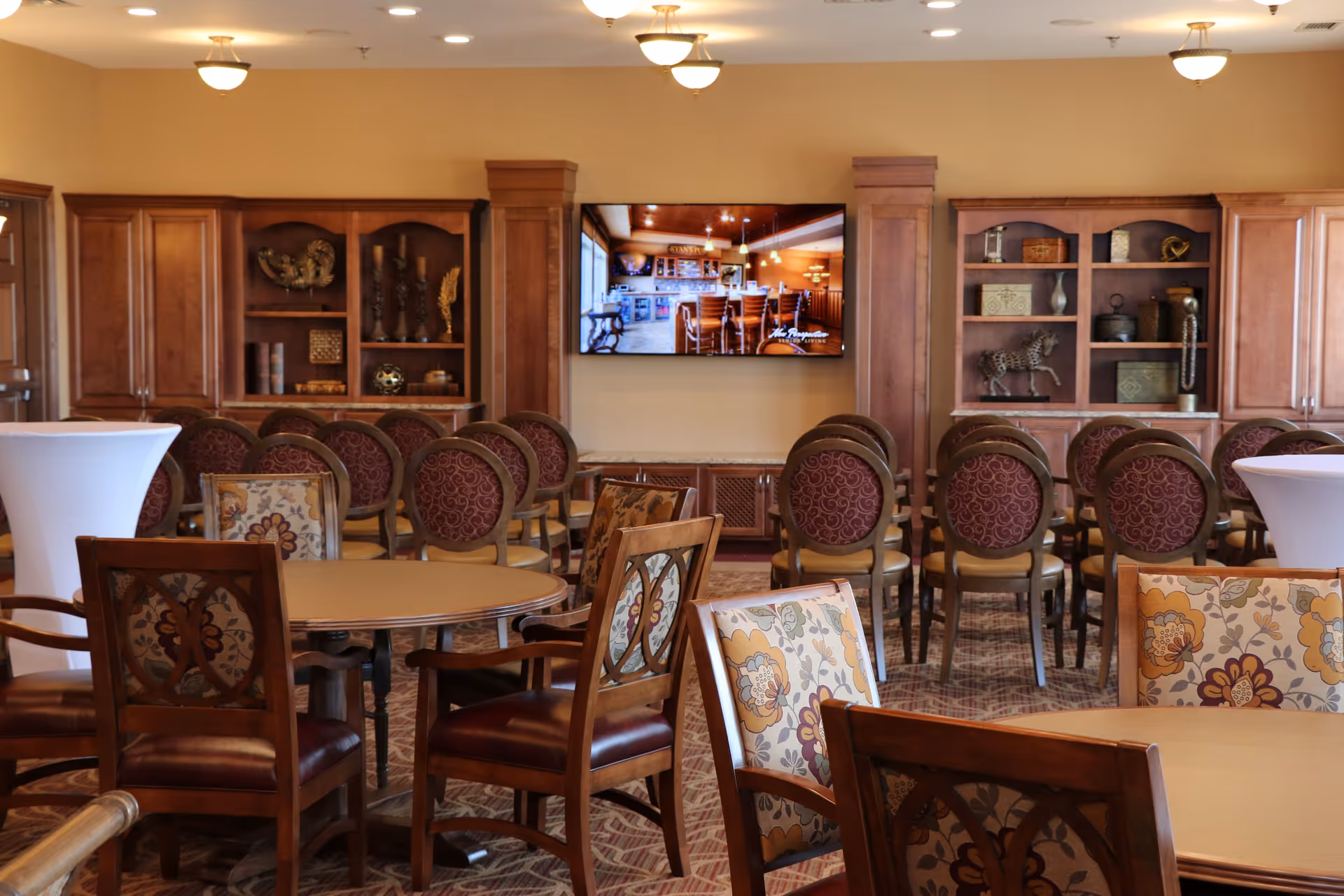 A well-lit senior living facility common area with multiple round tables and chairs arranged for seating. The chairs have floral and patterned upholstery. Wooden cabinets with decorative items line the back wall, and a flat-screen TV is mounted between two wooden columns. The carpet has a patterned design, and the ceiling has several light fixtures.