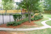Outdoor area with a curved concrete pathway, green grass, trees, shrubs, and a fenced section with a wooden shed in the background.