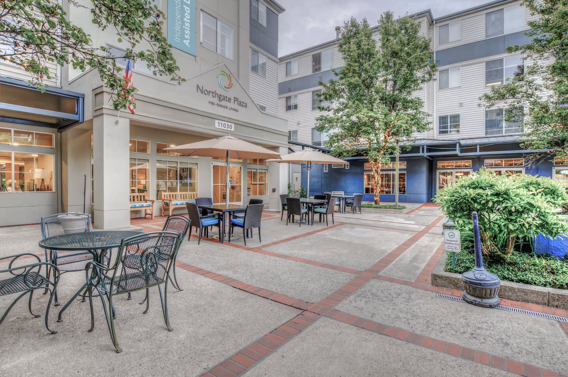 Outdoor courtyard area of Northgate Plaza senior living facility with metal tables and chairs, umbrellas, trees, and shrubs. The building exterior is visible with large windows and a sign that reads Northgate Plaza H&K Senior Living.