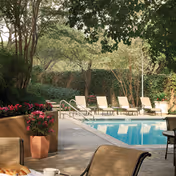 Outdoor swimming pool area with several lounge chairs arranged along the poolside, surrounded by trees and greenery. There are potted plants with pink flowers near the pool, and patio furniture including a table and chairs in the foreground.