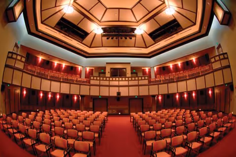 Wide-angle view of an empty auditorium with rows of chairs facing a stage and a two-tiered balcony beneath a domed ceiling.