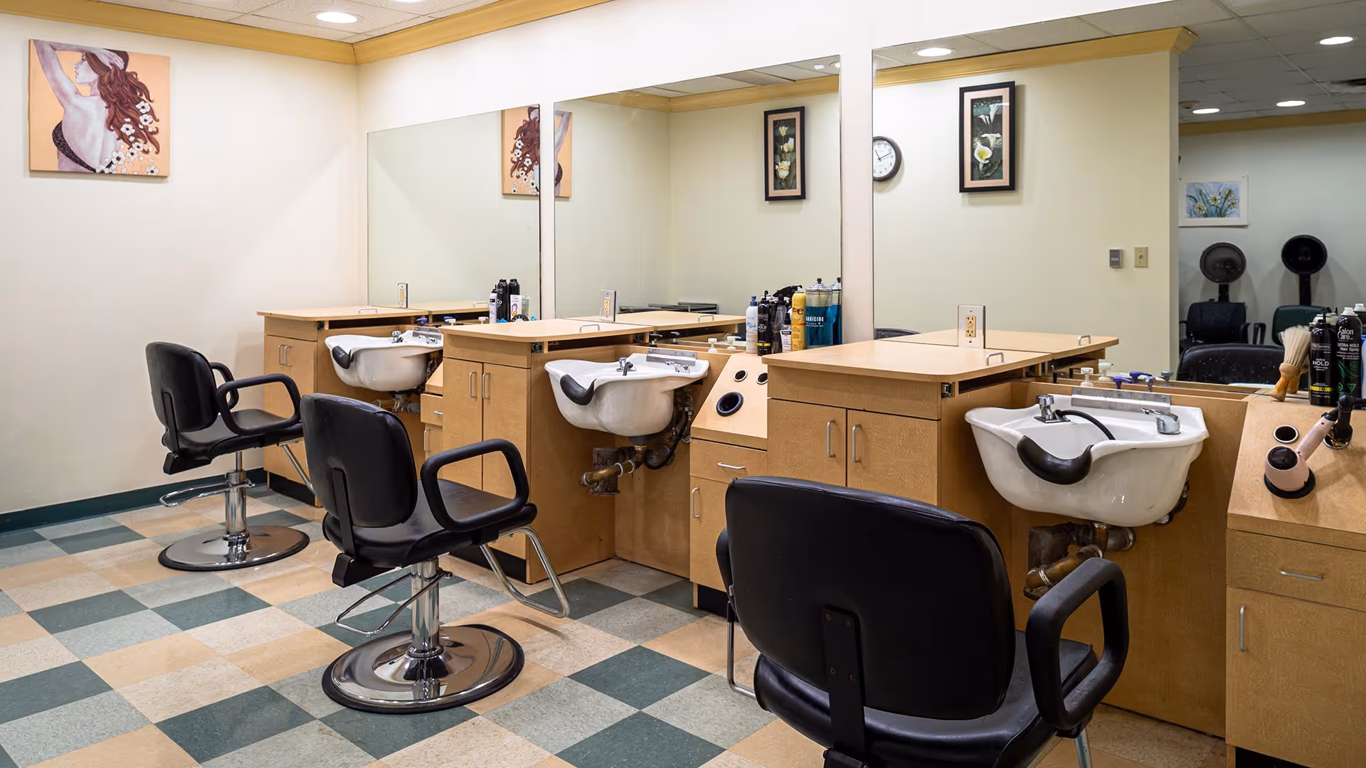Interior view of a hair salon area with three black salon chairs in front of wooden counters with built-in sinks. Large mirrors cover the walls behind the counters, and various hair care products are placed on the counters. The floor has a checkered pattern with green and beige tiles, and framed artwork decorates the walls.