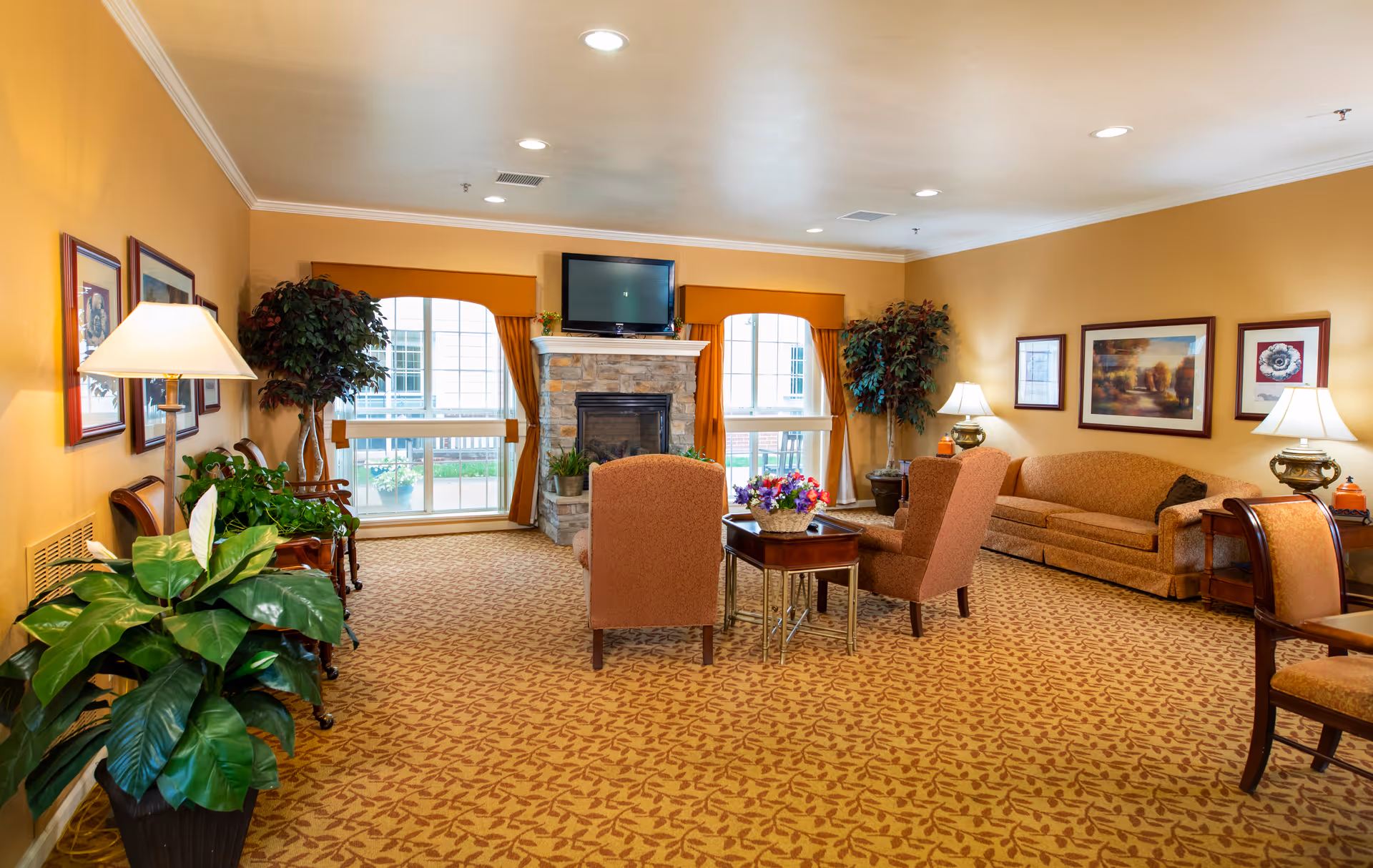 A cozy living room in an assisted living facility featuring a stone fireplace with a TV mounted above it, two large windows with orange curtains, several upholstered chairs and a sofa, framed artwork on the walls, table lamps, and green potted plants. The room has warm yellow walls and patterned carpet flooring.
