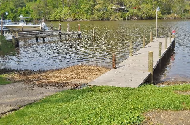 A wooden dock extending over a calm body of water with green grass and a small sandy area in the foreground. Trees and additional docks are visible across the water.