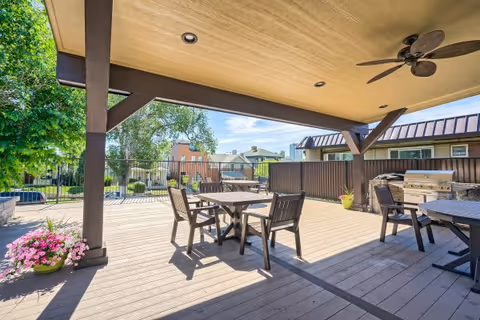 Covered outdoor patio area with wooden deck flooring, several tables and chairs, ceiling fan, and a barbecue grill. There are potted plants and a fenced garden area with trees and shrubs in the background under a clear blue sky.